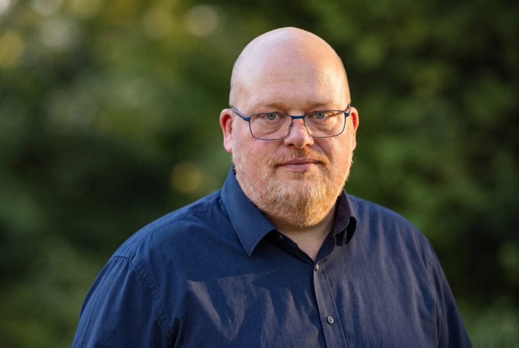 Porträt eines Mannes mit Brille und dunkelblauem Hemd vor unscharfem grünen Hintergrund